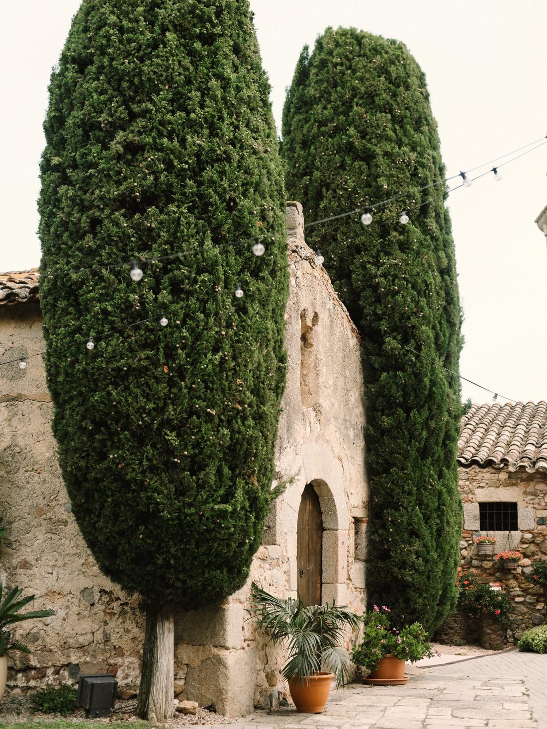 Cypresses flanking the chapel door of a stone quinta, festoon lights strung overhead